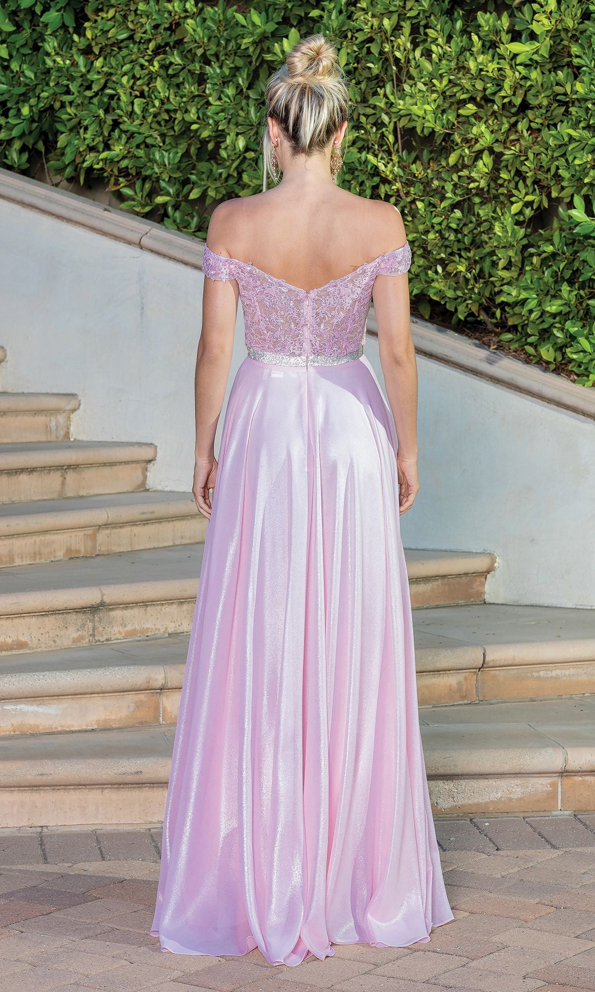 A blonde woman with her hair in a bun stands outside, facing away, wearing the Sheer Embroidered-Bodice Metallic Long Prom Dress—an off-the-shoulder, floor-length gown with a lace bodice and flowing skirt.